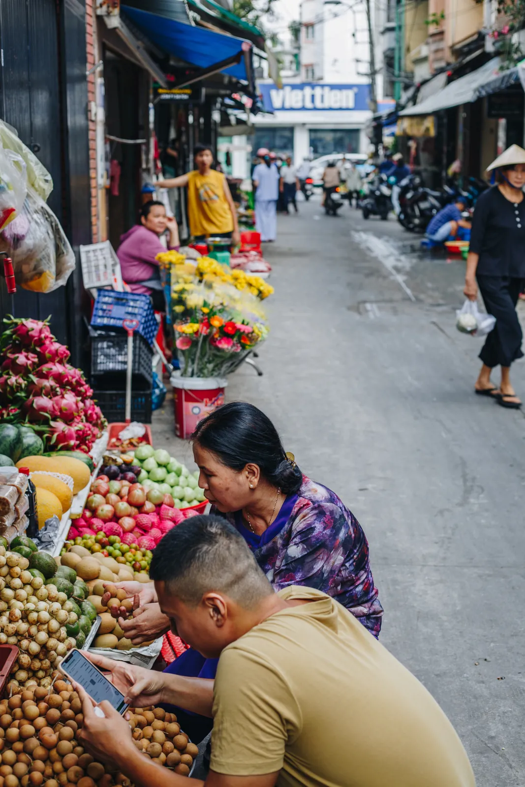 Two guests examining fruit at a crowded market stall on a Saigon street