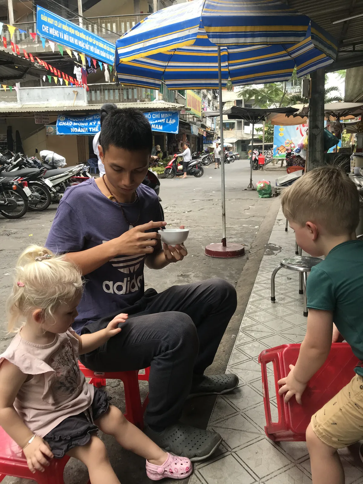 Two children sitting with a man eating on plastic stools on a busy street